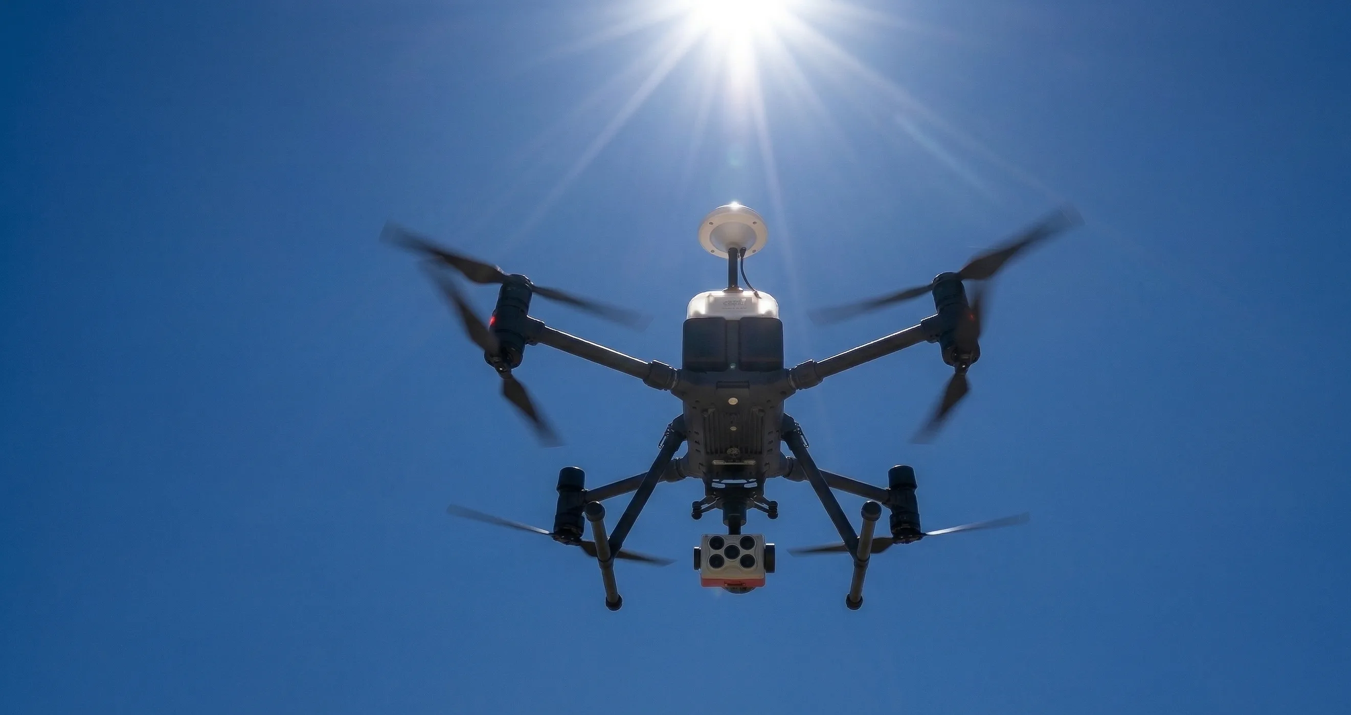Underside view of a drone carrying a specialty sensor payload under strong midday sun.